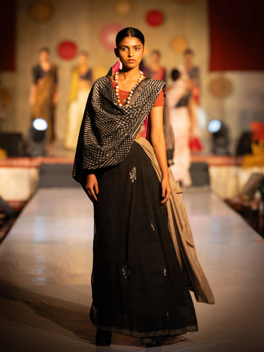 Model walking on a fashion show runway wearing a black saree with a patterned shawl. Bani Thani Chandrika Saree by Soulweaves