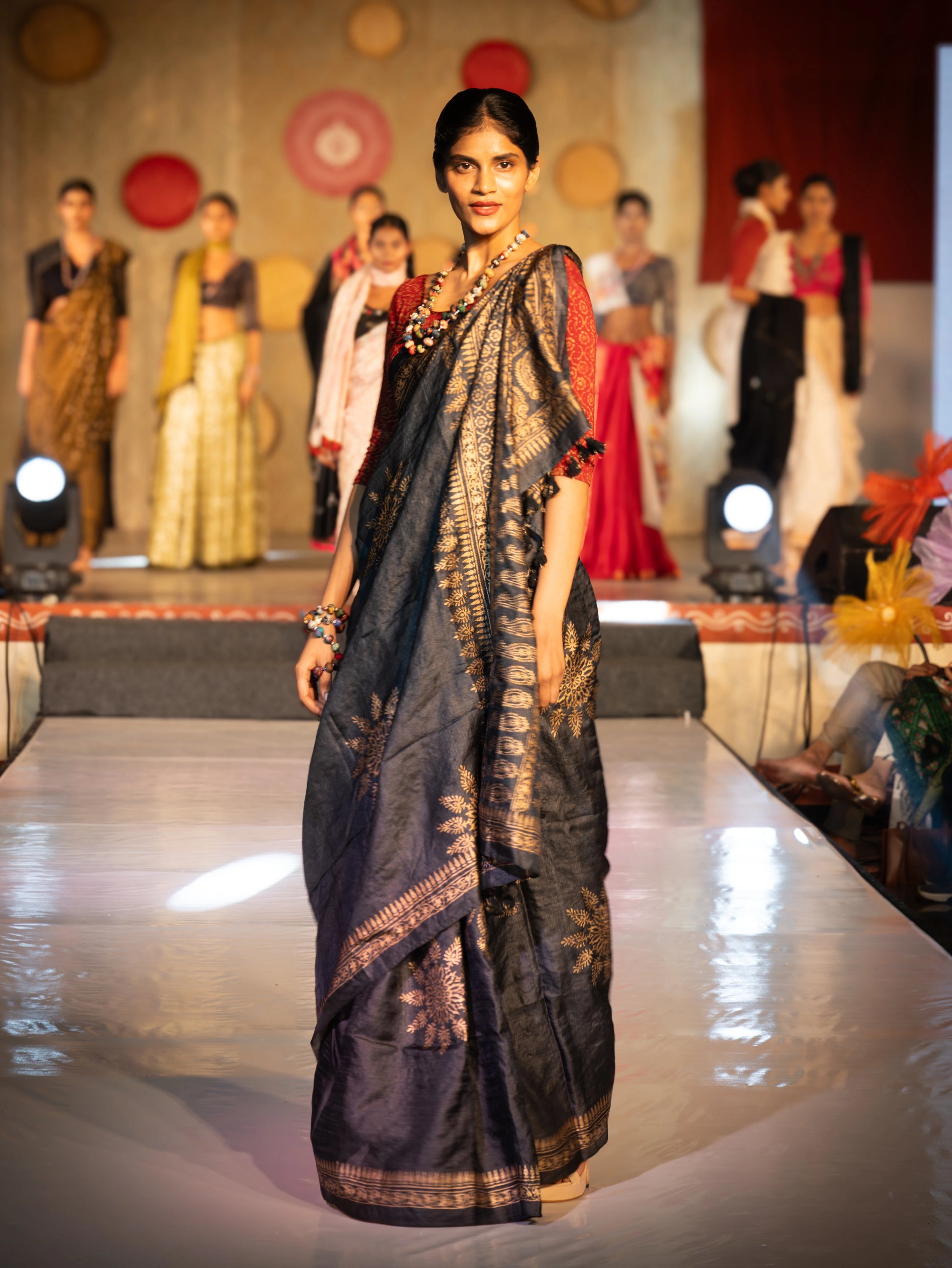 Woman in a saree walking on a fashion show runway with other models in the background. Bani Thani Rajsi Saree by Soulweaves